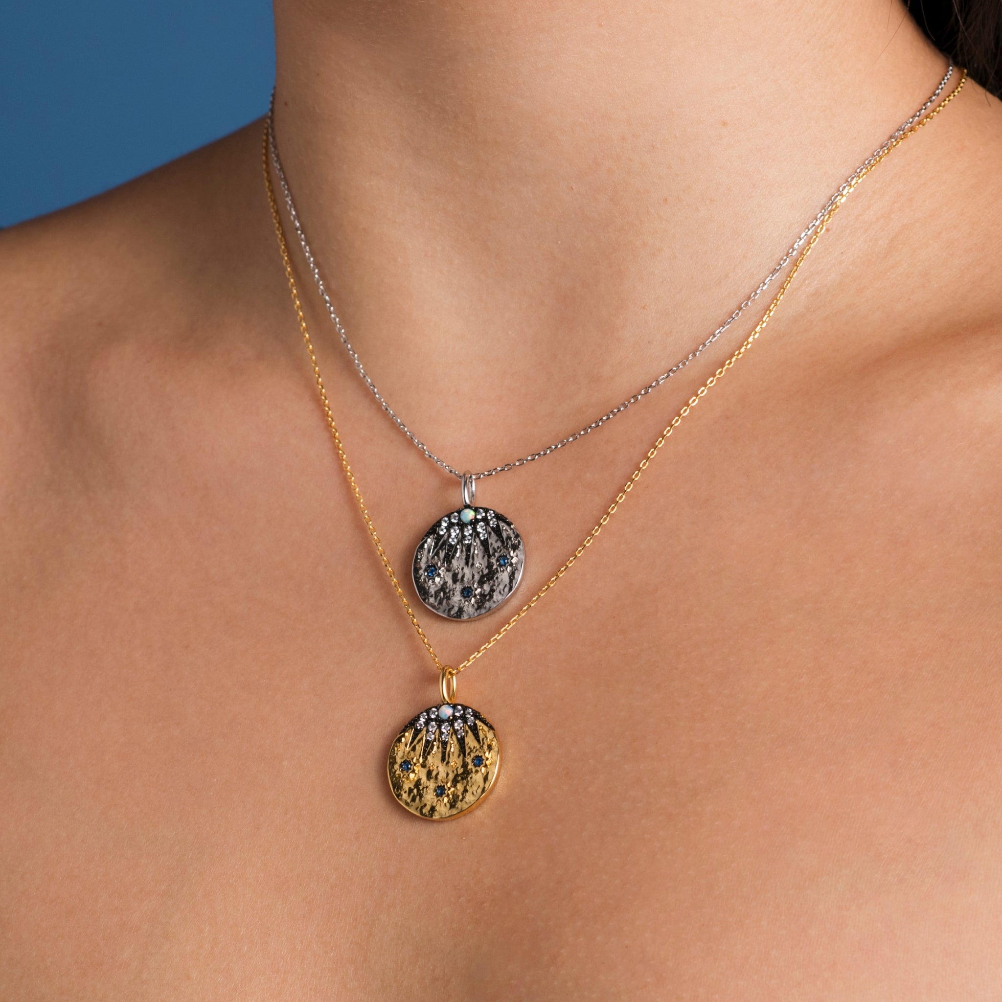 A woman wearing a layered necklace with two textured pendants, one featuring a sapphire blue cubic zirconia, against a blue background.