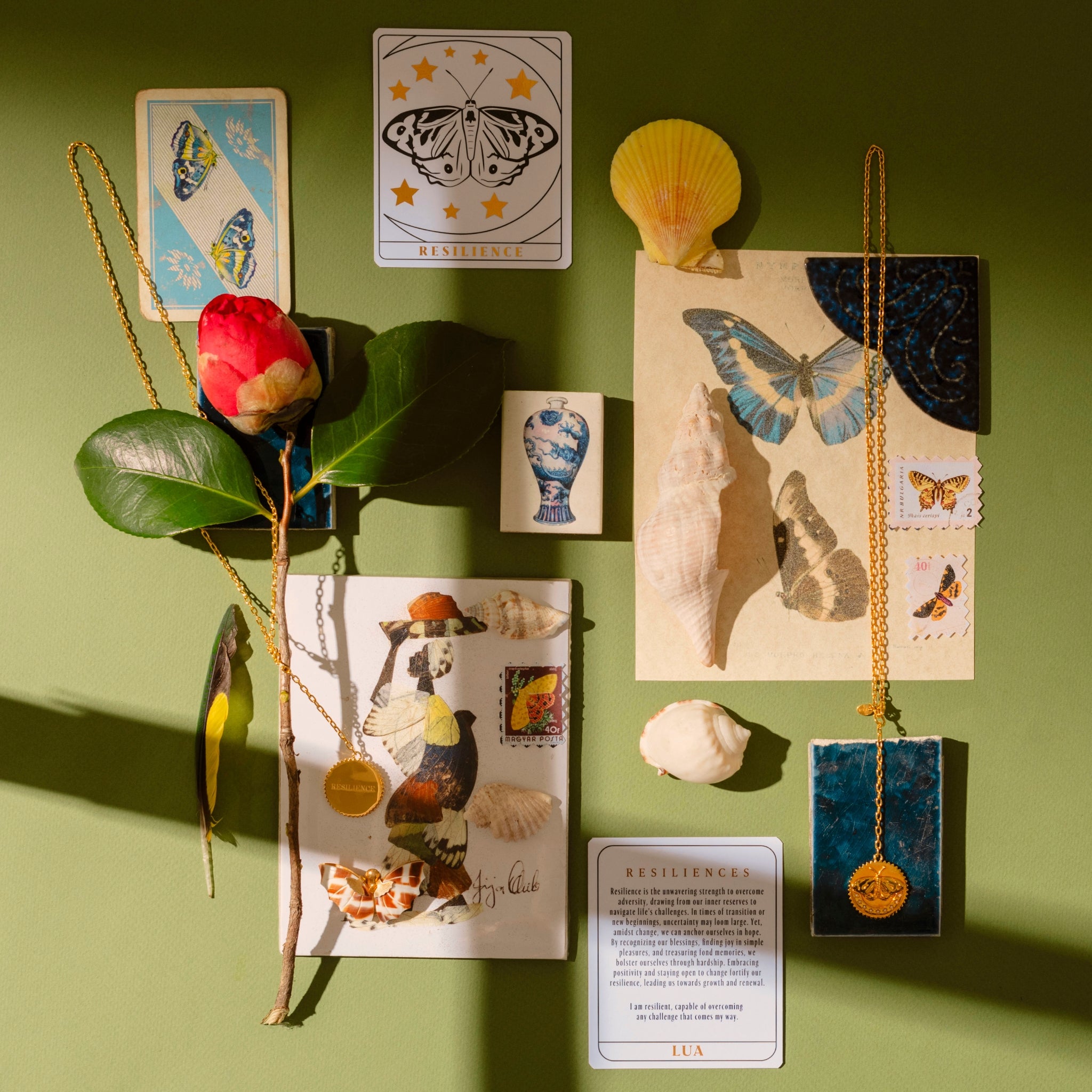 An arrangement of various objects including seashells, butterfly illustrations, a red flower, a tarot card symbolizing transformation, two gold necklaces, and decorative cards on a green surface.