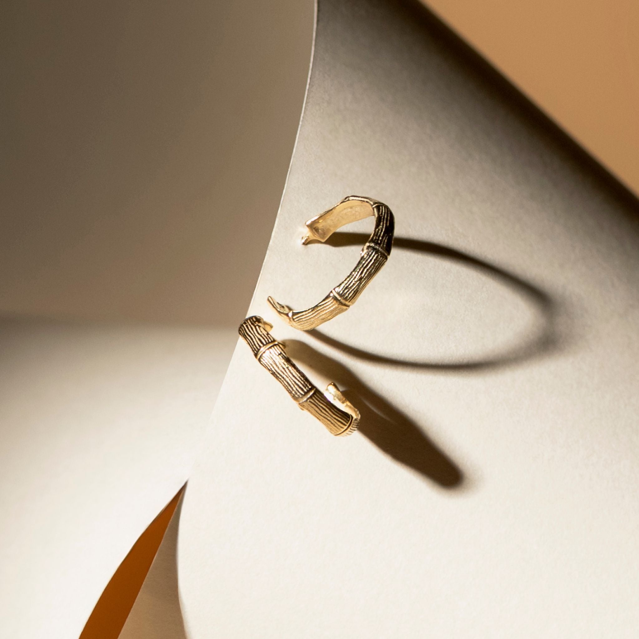 A pair of gold-toned bamboo-style hoop earrings with textured grooved details, displayed on a curved cream-colored surface with dramatic shadows.