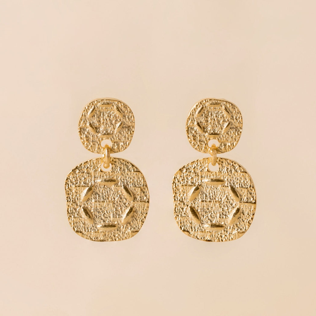 A pair of gold textured drop earrings with a small circular stud and a larger rounded square pendant, shown on a solid pale beige background.