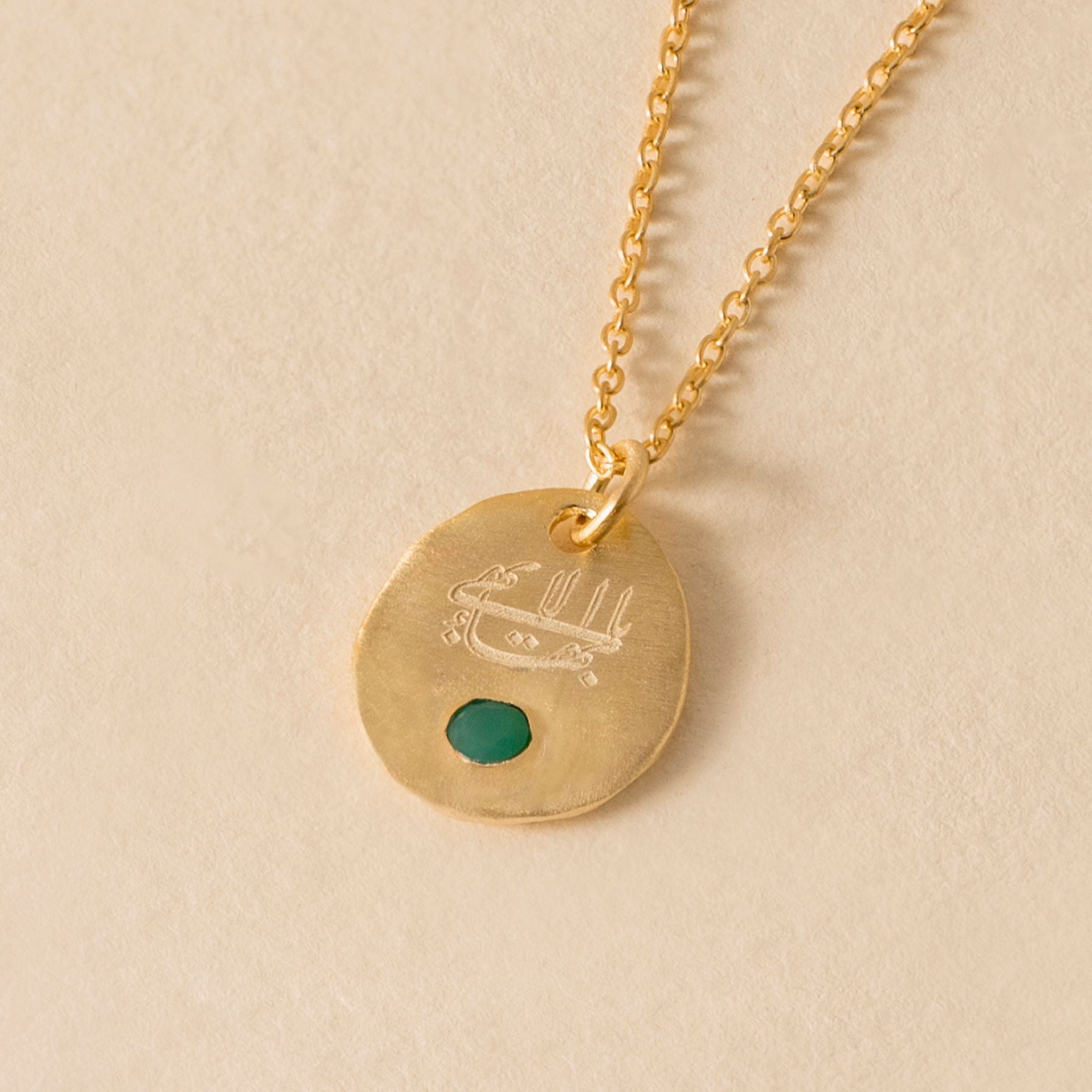 Gold necklace with an oval pendant featuring the greatest name calligraphy and a green stone on a beige background