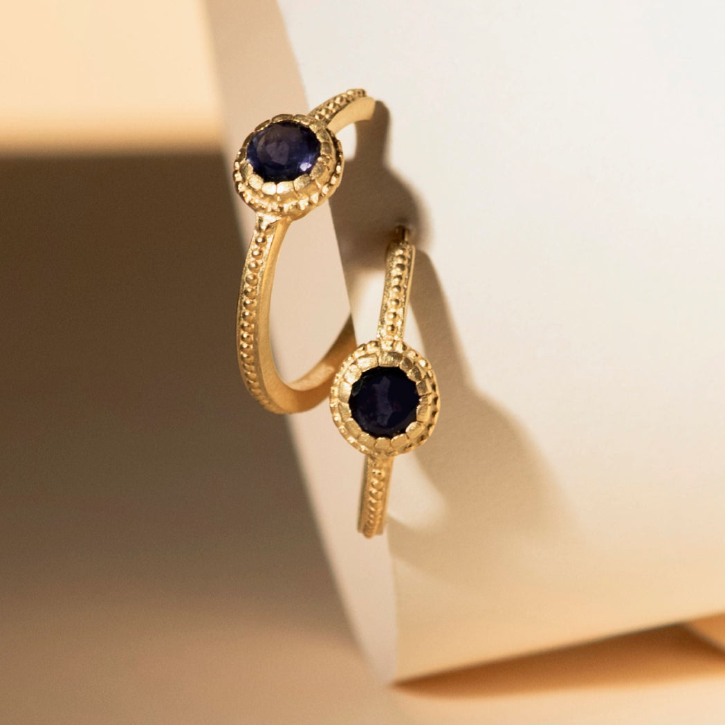 Two gold rings with oval dark blue gemstones and textured halo settings, displayed on a cream-colored jewelry stand.