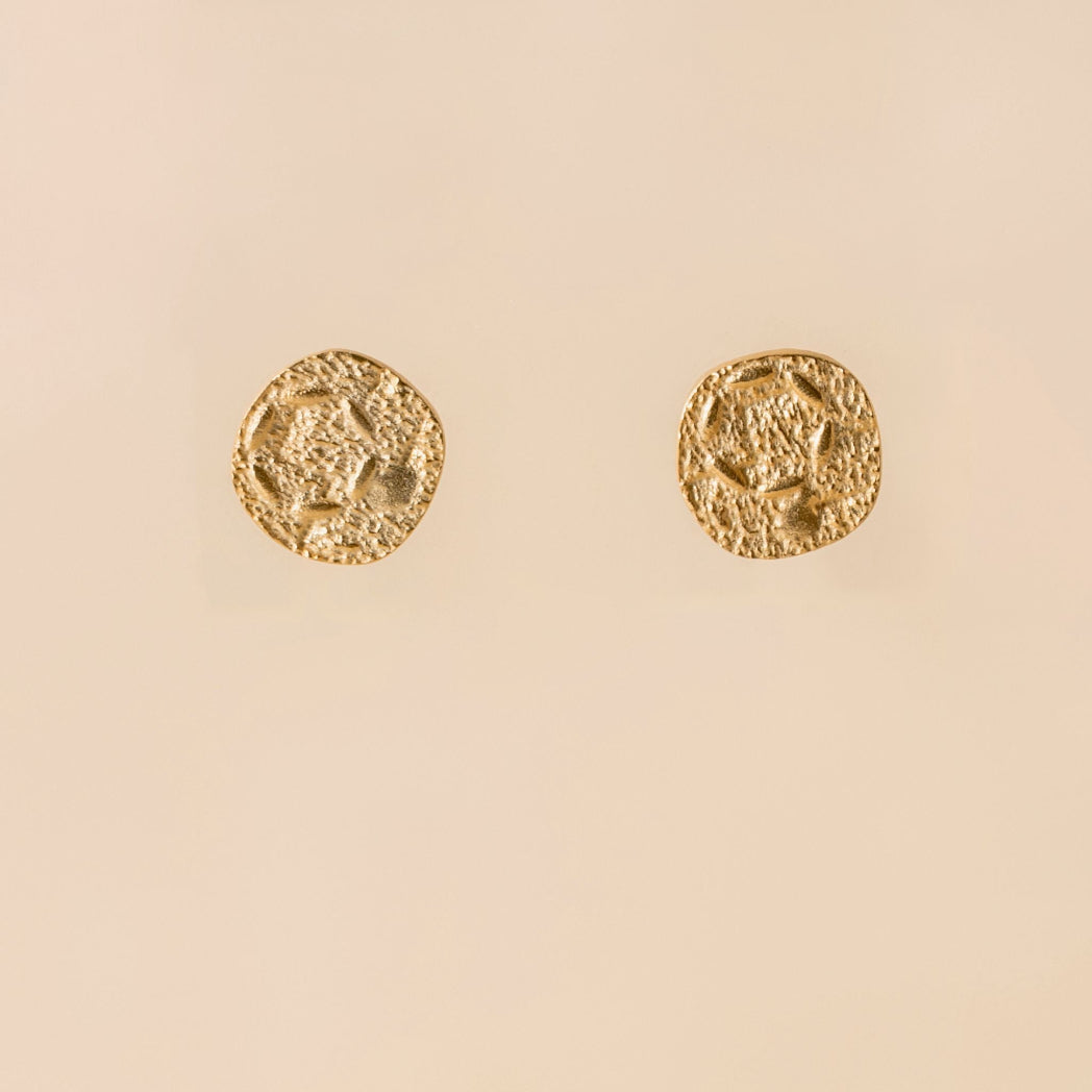 A pair of textured gold coin-shaped stud earrings on a solid pale peach-beige background.