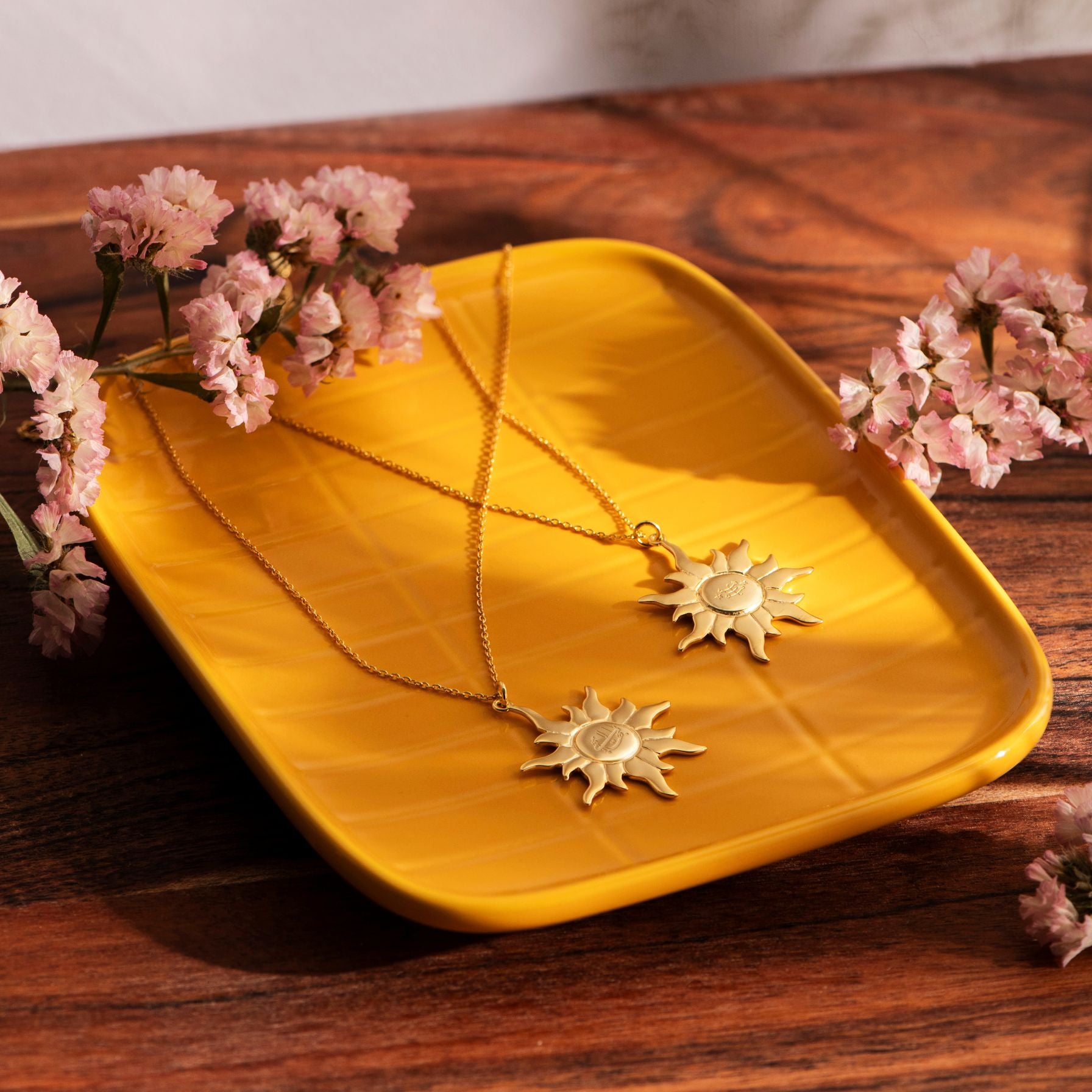Gold sun-shaped jewelry on a yellow tray with pink flowers on a wooden surface