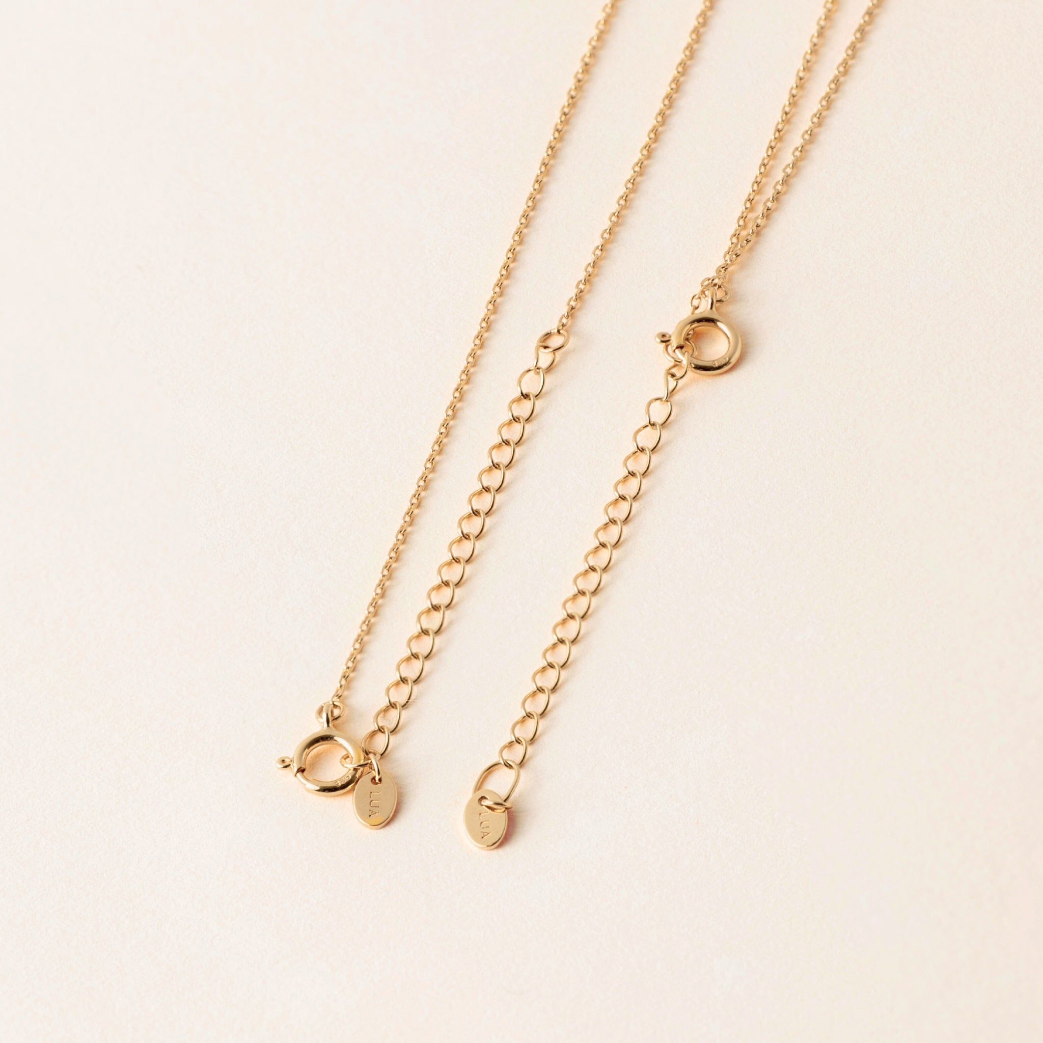 Two gold chains with delicate extension chains for adjustable length laid on a light beige surface, each featuring small circular clasps and bespoke enamel pieces.