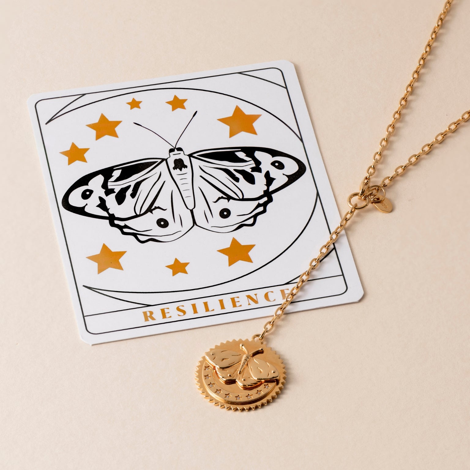 A gold Resilience Butterfly Necklace lies on a card featuring a black-and-white butterfly with surrounding gold stars and the word "RESILIENCE" at the bottom, symbolizing unwavering spirit and transformation.