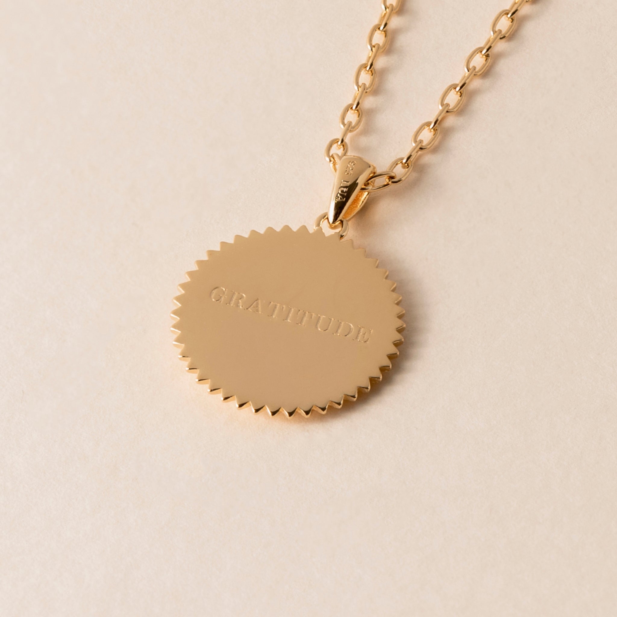 The Gratitude Hummingbird Necklace features a gold necklace with a round pendant inscribed with the word "GRATITUDE." The pendant, accentuated by a serrated edge design, is a beautiful reminder of life's blessings.