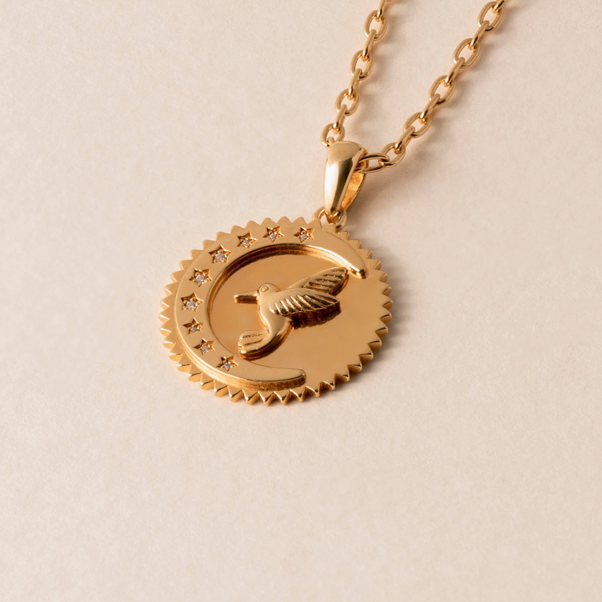 Close-up of a gold Gratitude Hummingbird Necklace featuring a pendant with a bird and seven stars within a circular design, symbolizing life's blessings.