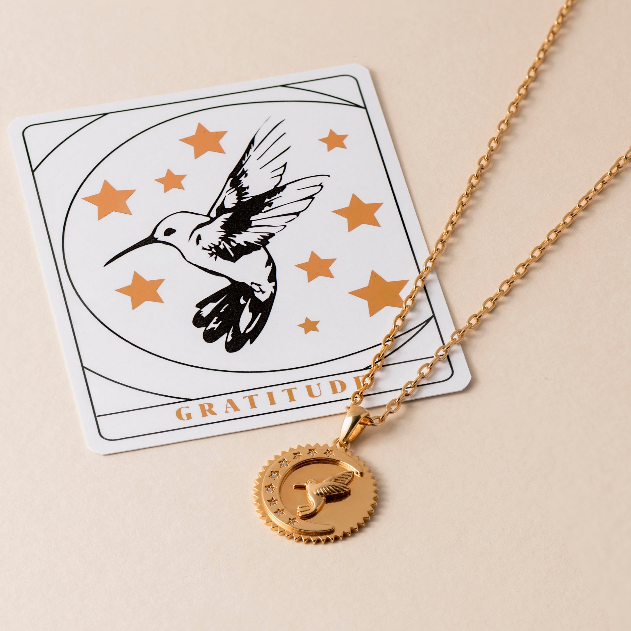 A gold necklace with a pendant featuring a hummingbird, symbolizing life's blessings, lies on a beige surface. Behind it is a card with a similar hummingbird and star design, along with the word "GRATITUDE" printed on it. This Gratitude Hummingbird Necklace celebrates well-being and growth.