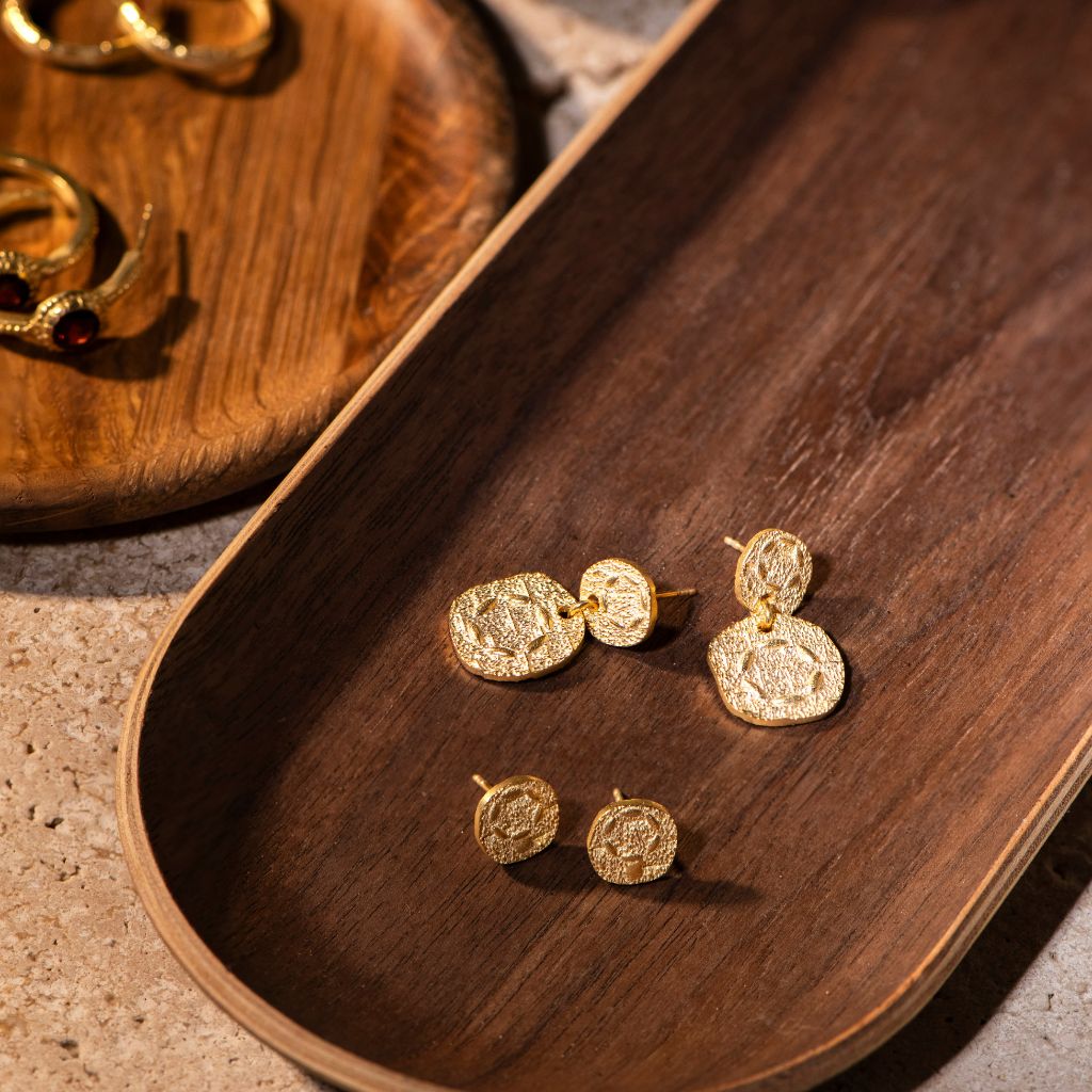 Gold coin drop earrings with textured, embossed details, displayed on a dark wooden tray.