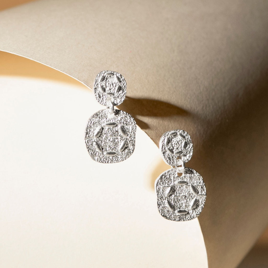 A pair of silver-toned double-drop statement earrings with textured pavé detailing, displayed on a beige jewelry stand.