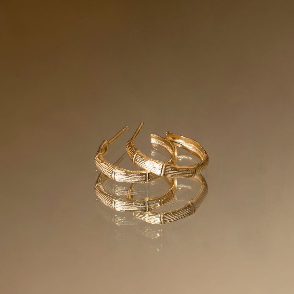 A pair of textured gold hoop earrings with a ribbed, bamboo-like pattern, displayed on a reflective surface against a warm brown gradient background.