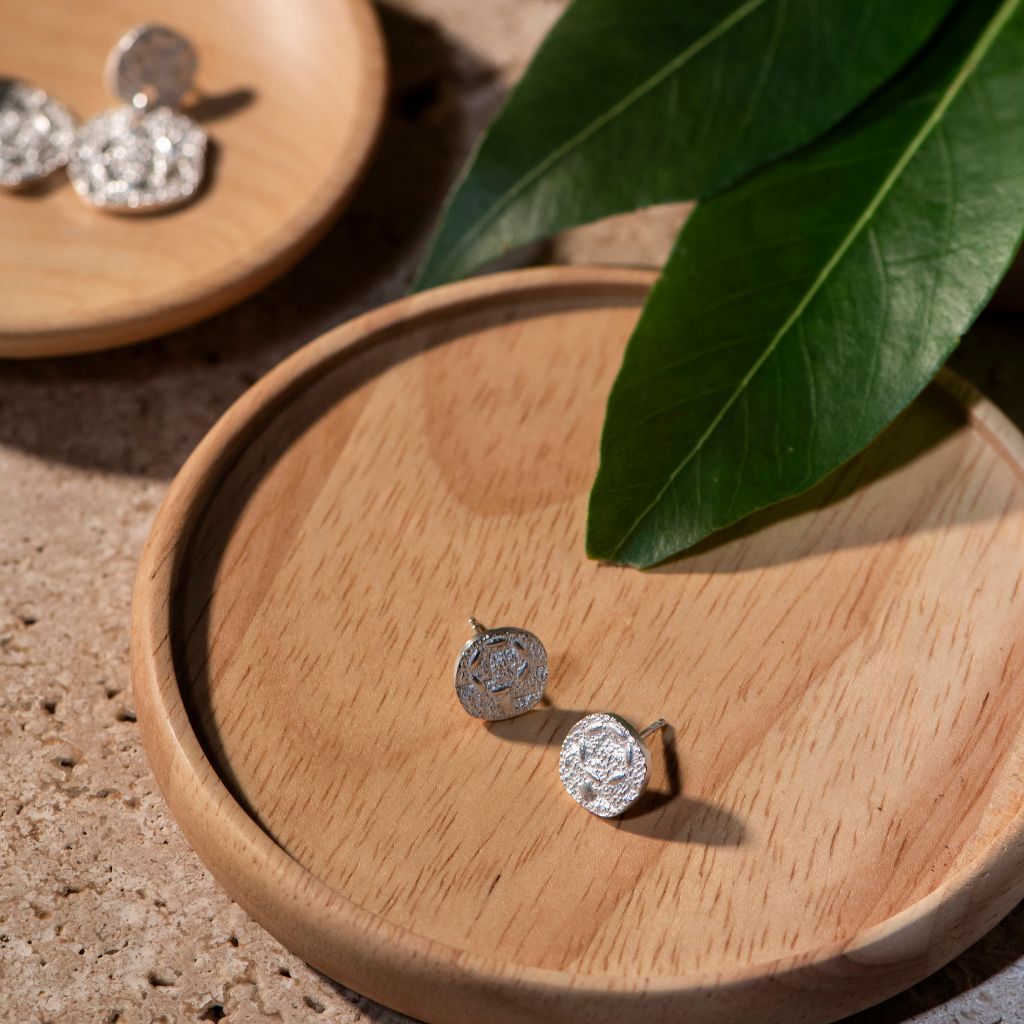 Two silver coin-style stud earrings displayed on a round wooden tray, with additional earrings on another tray in the background and a green leaf in the foreground.