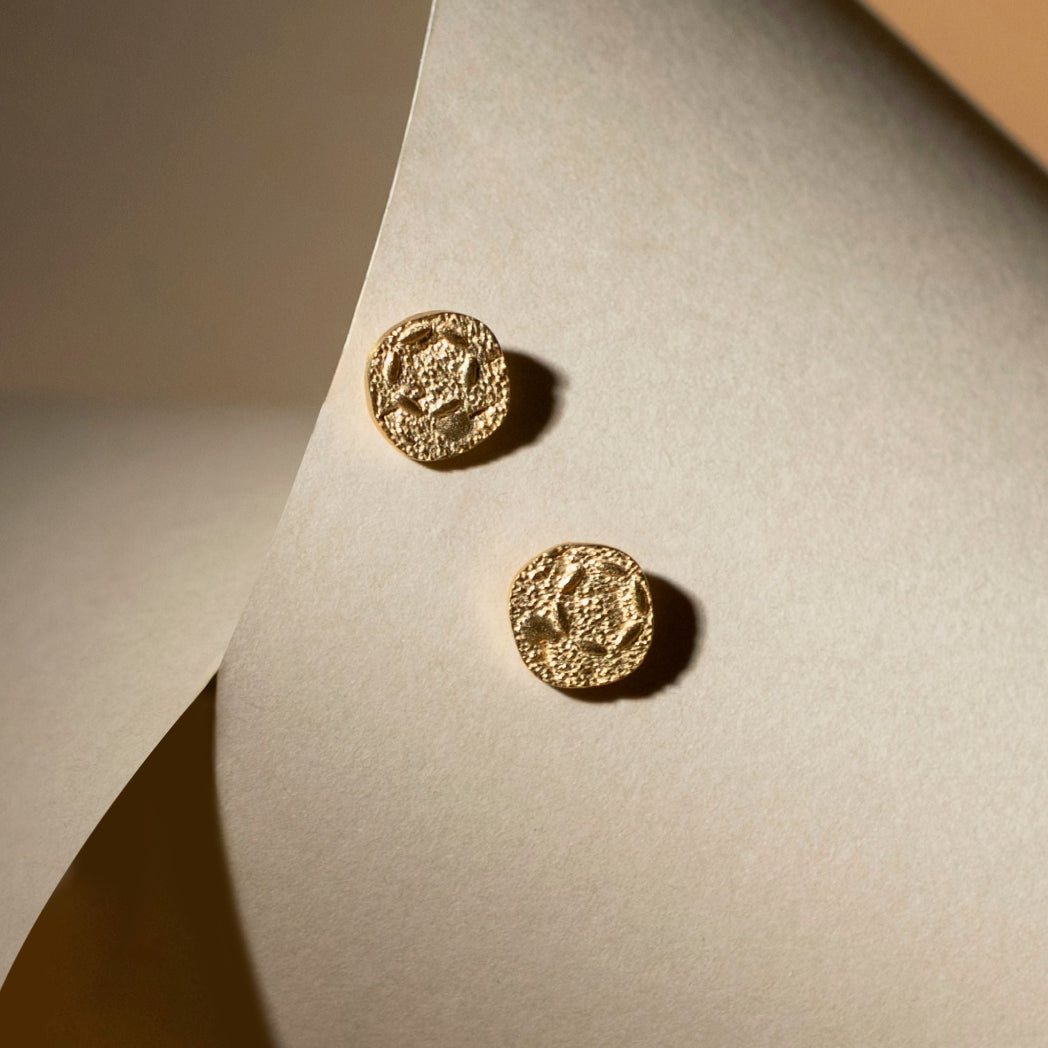 A pair of textured gold round stud earrings displayed on a curved beige paper surface with warm directional lighting.