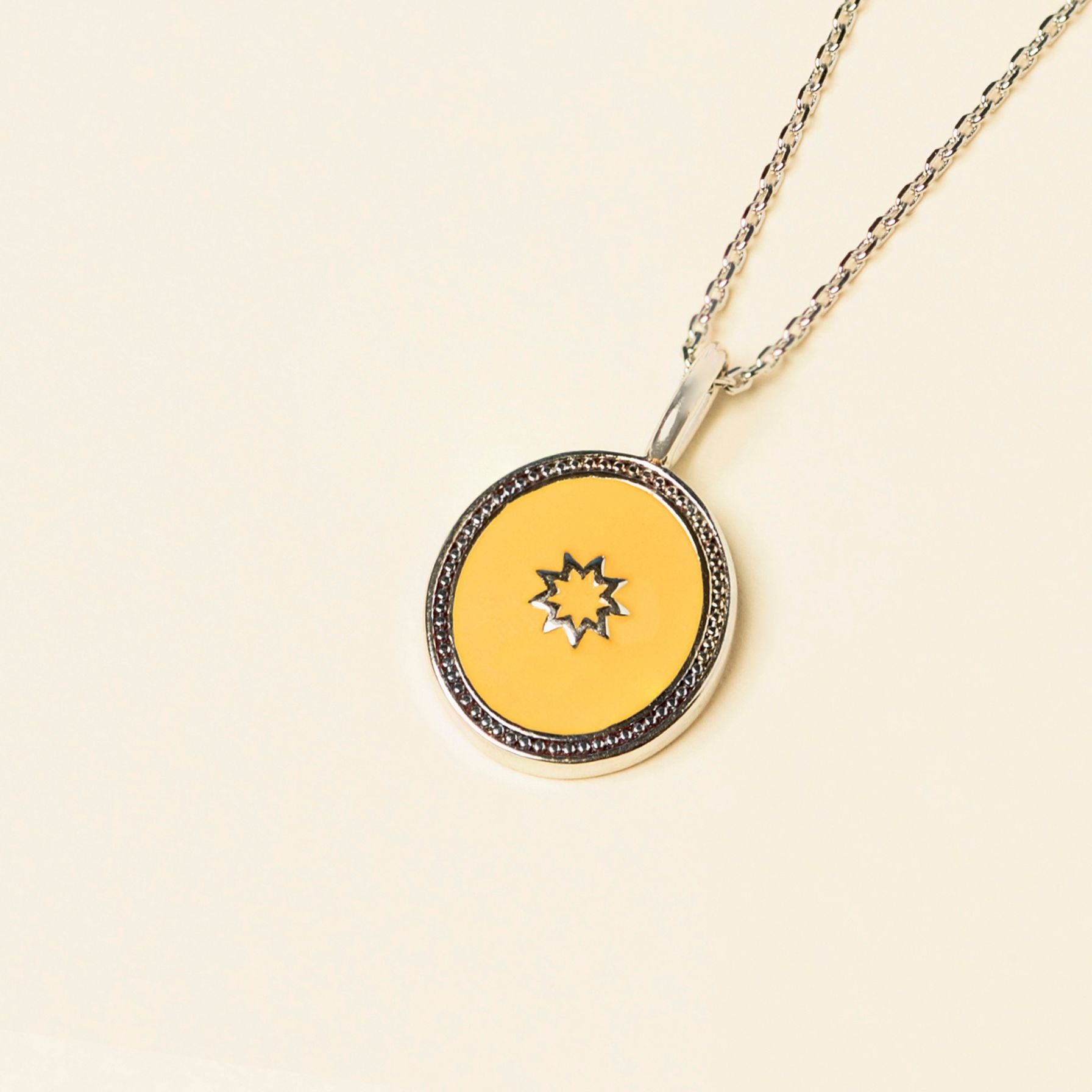 A pendant with a bespoke enamel center featuring a small starburst design, surrounded by a silver frame, on a silver chain against a cream background.