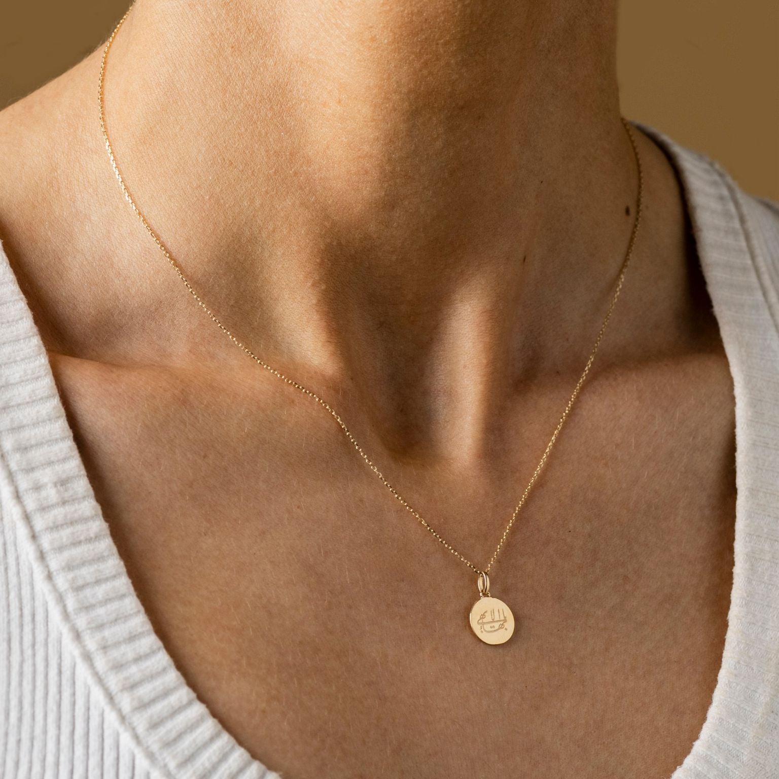 Close-up of a person wearing a fine gold necklace with a round gold pendant from the Lua collection that has an inscriptionof the Ringstone Symbol. The person is dressed in a white ribbed top.