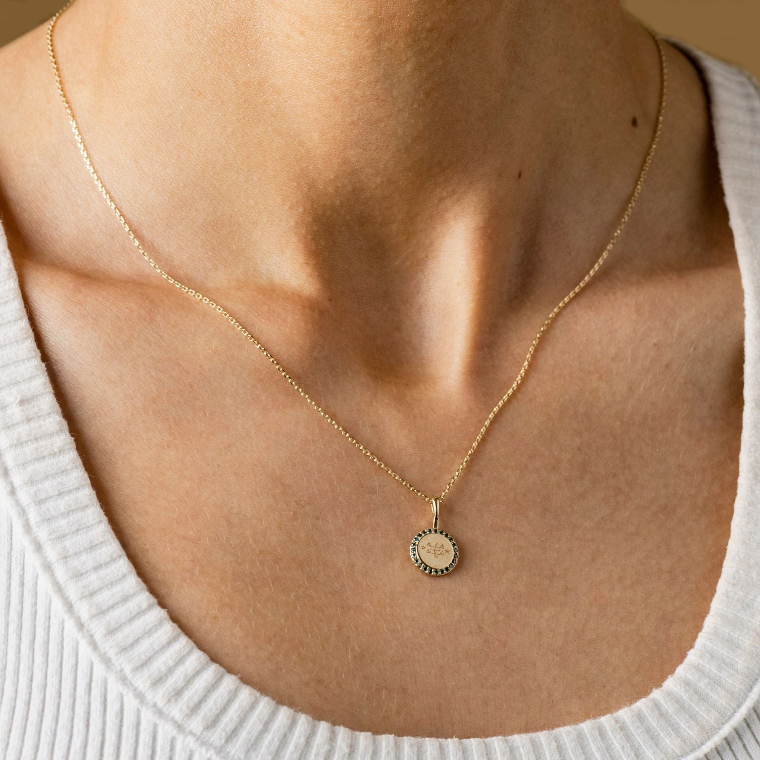 A person wearing a gold necklace with a round pendant featuring an engraved Ringstone symbol, encircled by blue diamonds