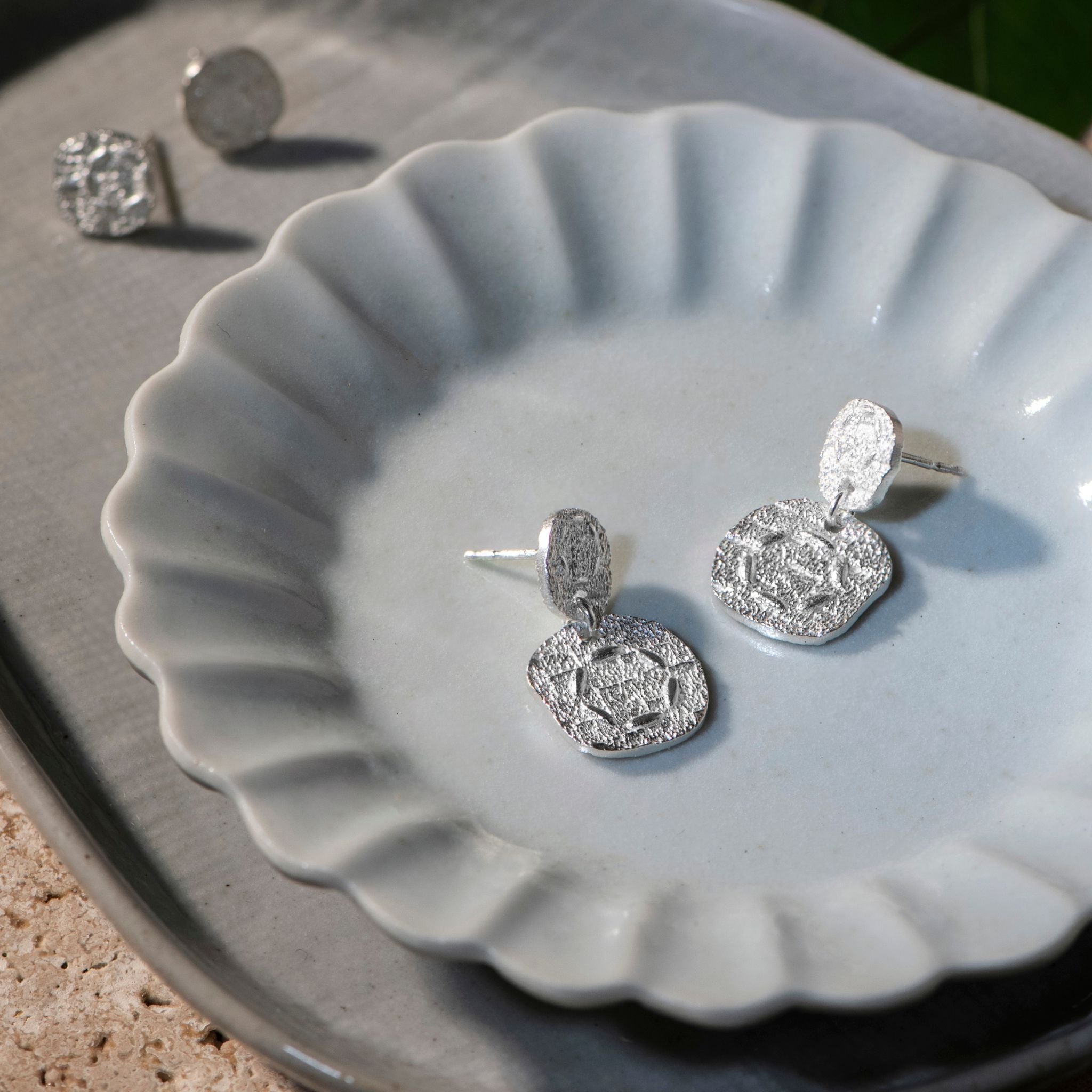 Silver textured coin drop earrings displayed on a light gray scalloped ceramic dish, with a pair of matching silver stud earrings resting on the gray surface in the background.