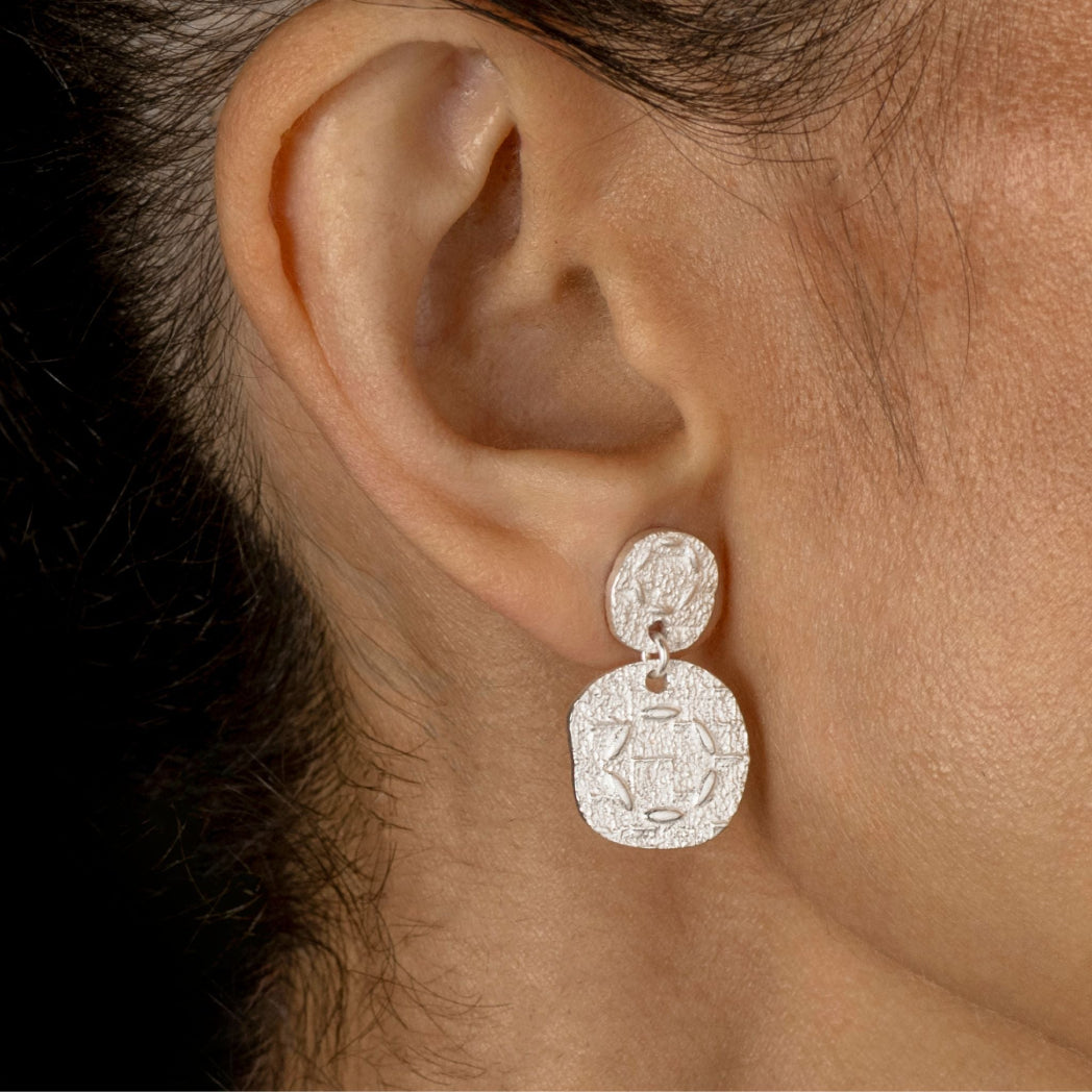 A close-up of a person's ear wearing a silver dangle earring with two textured oval charms.