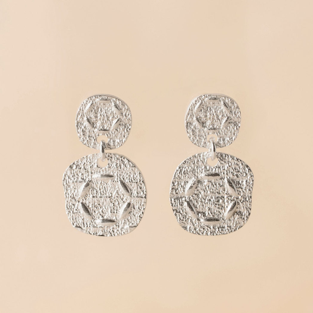 A pair of textured silver drop earrings with circular and geometric dangle designs, displayed against a solid beige background.