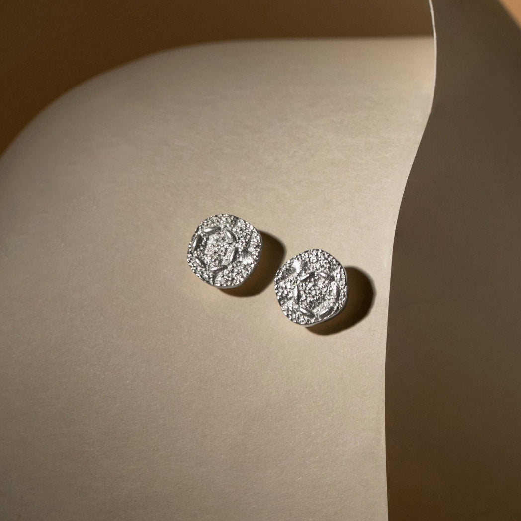 A pair of round silver stud earrings with textured, oxidized details, displayed on a beige curved surface.