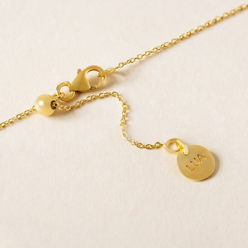Gold necklace clasp detaill with a pearl and an amethyst pendant engraved with the name "Lua" on a textured cream background.
