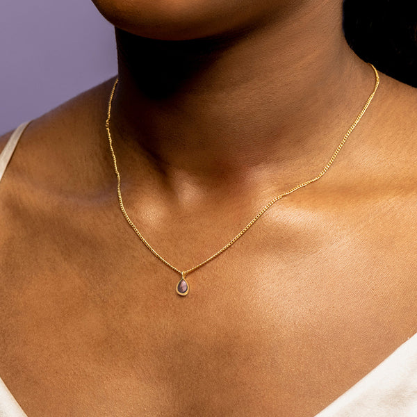 Close-up of a woman's neck wearing a delicate gold necklace with a small amethyst pendant, against a purple background.