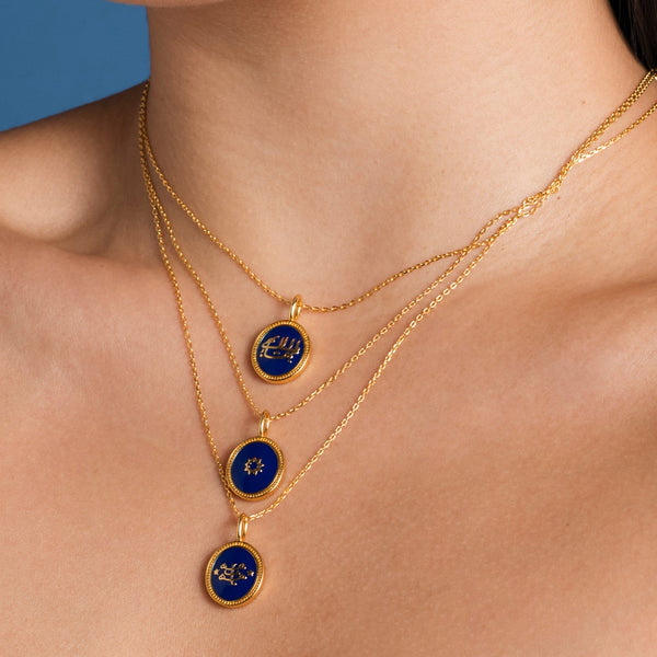 Close-up of a woman wearing a layered gold necklace with three blue Bahai Faith symbols pendant charms against a neutral background.