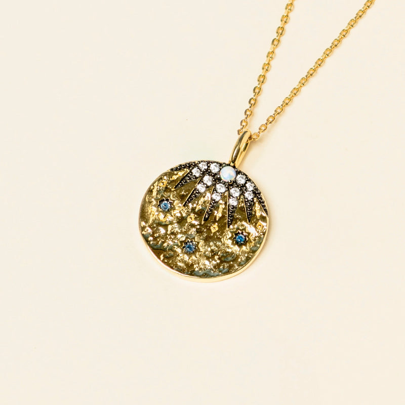 Gold pendant necklace with sapphire blue cubic zirconia accents and starburst design on a beige background.