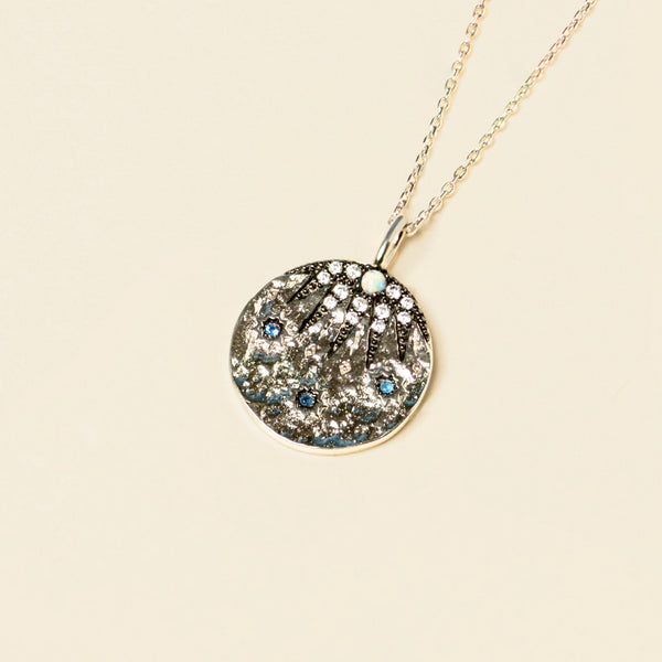 A pendant necklace with a circular, tree-themed charm embellished with sapphire blue cubic zirconia on a light background.