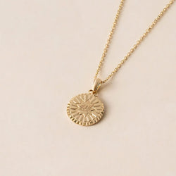 A gold necklace with a round, textured pendant on a thin chain, featuring an engraving of the greatest name sybol inside a nine pointed star displayed on a beige background.