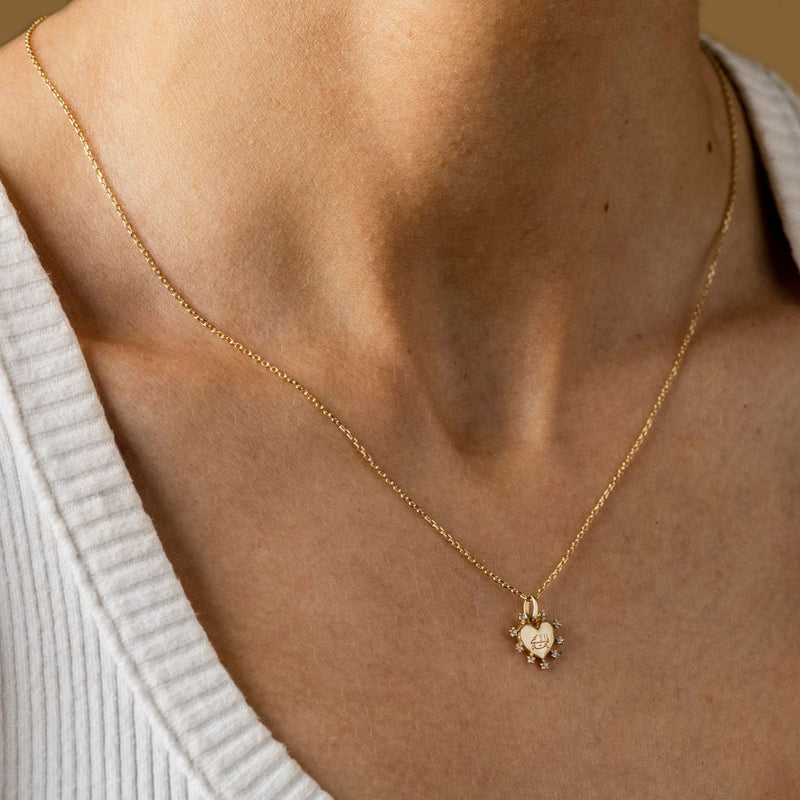 A person wearing a Gold heart-shaped pendant necklace with small cubic zirconia gemstones outlining the heart and featuring a subtle Greatest Name engraving