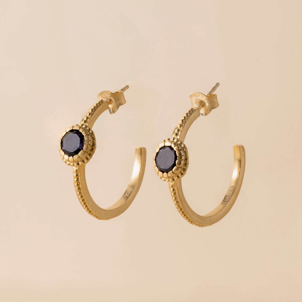 A pair of gold huggie hoop earrings with textured beaded edges and round dark gray gemstones, shown on a solid beige background.