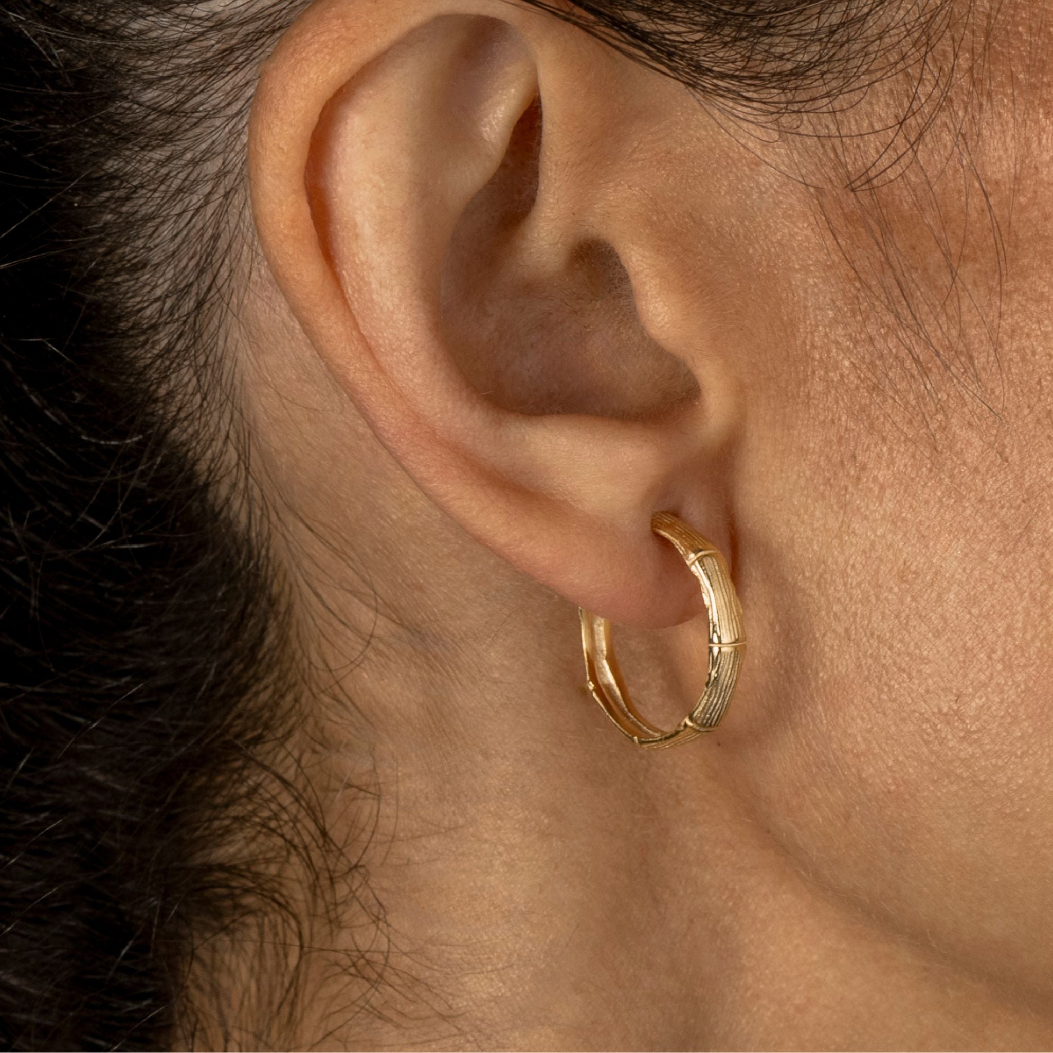 A close-up of a person's ear wearing a small gold hoop earring with a textured, bamboo-like design.