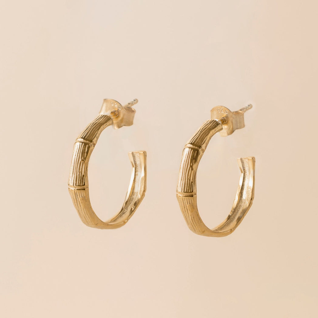 A pair of gold hoop earrings with a textured, segmented bamboo-inspired design, shown against a solid light beige background.