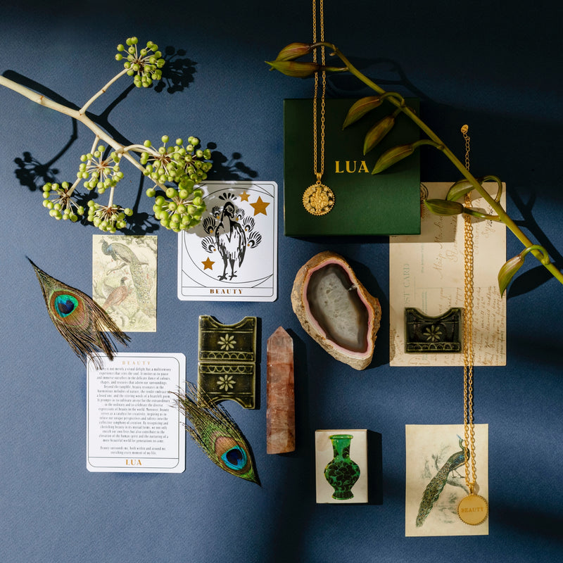 A flat lay of various objects including tarot cards, a Peacock Necklace, peacock feathers, a crystal, a geode, and a green box with a gold emblem, all arranged on a dark blue surface with plant branches. The arrangement offers rich sensory experiences and visual beauty.