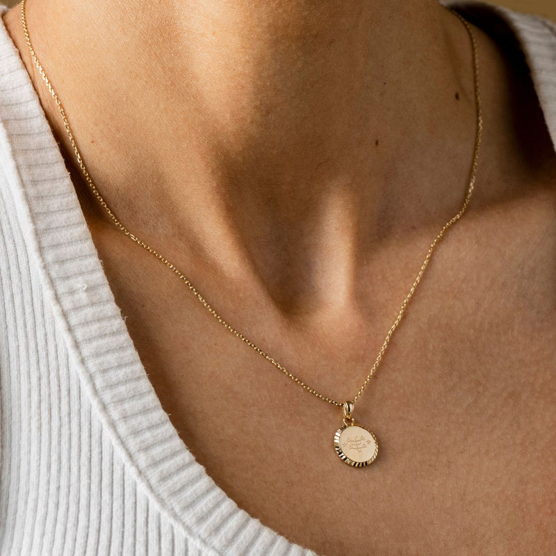 A person wearing a gold necklace with a round pendant featuring an engraved Ringstone Symbol design.