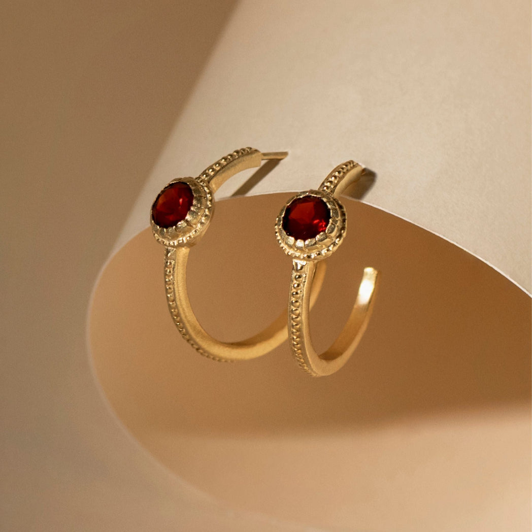 A pair of gold-toned hoop earrings with oval red gemstones and small clear stones around the settings, displayed on a beige jewelry stand.
