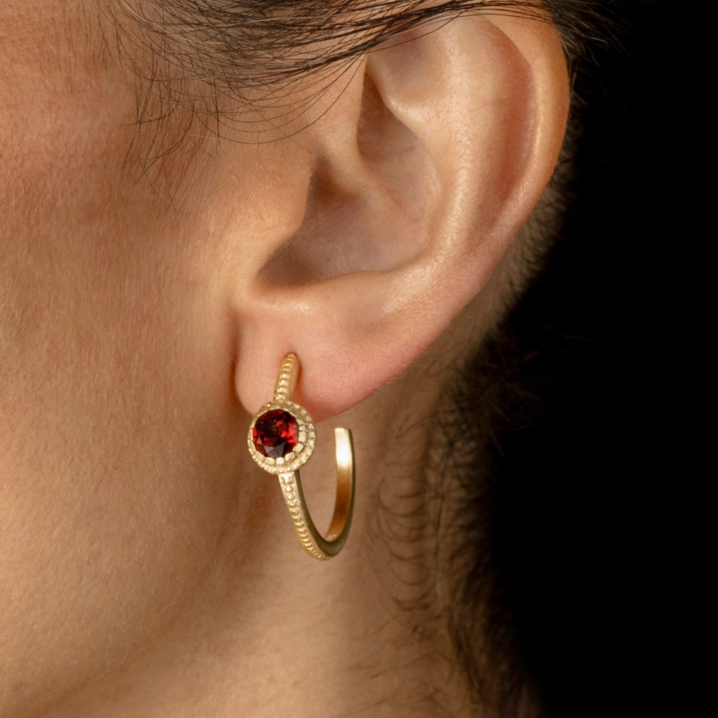A close-up of a person's ear wearing a gold hoop earring with a central round red gemstone surrounded by a halo of small clear stones.