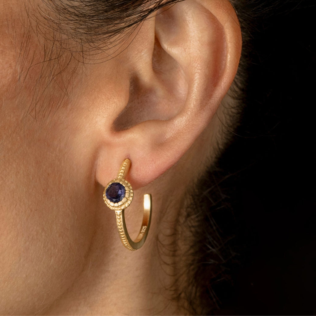 A close-up of a person's ear wearing a gold huggie hoop earring with a round dark blue gemstone surrounded by a halo of small clear stones.