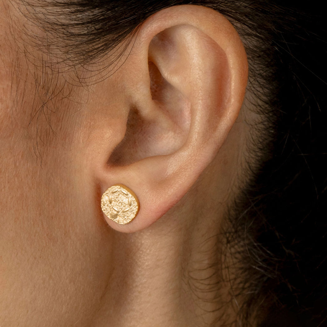 A close-up of a person's ear wearing a small round gold stud earring with an embossed textured design.