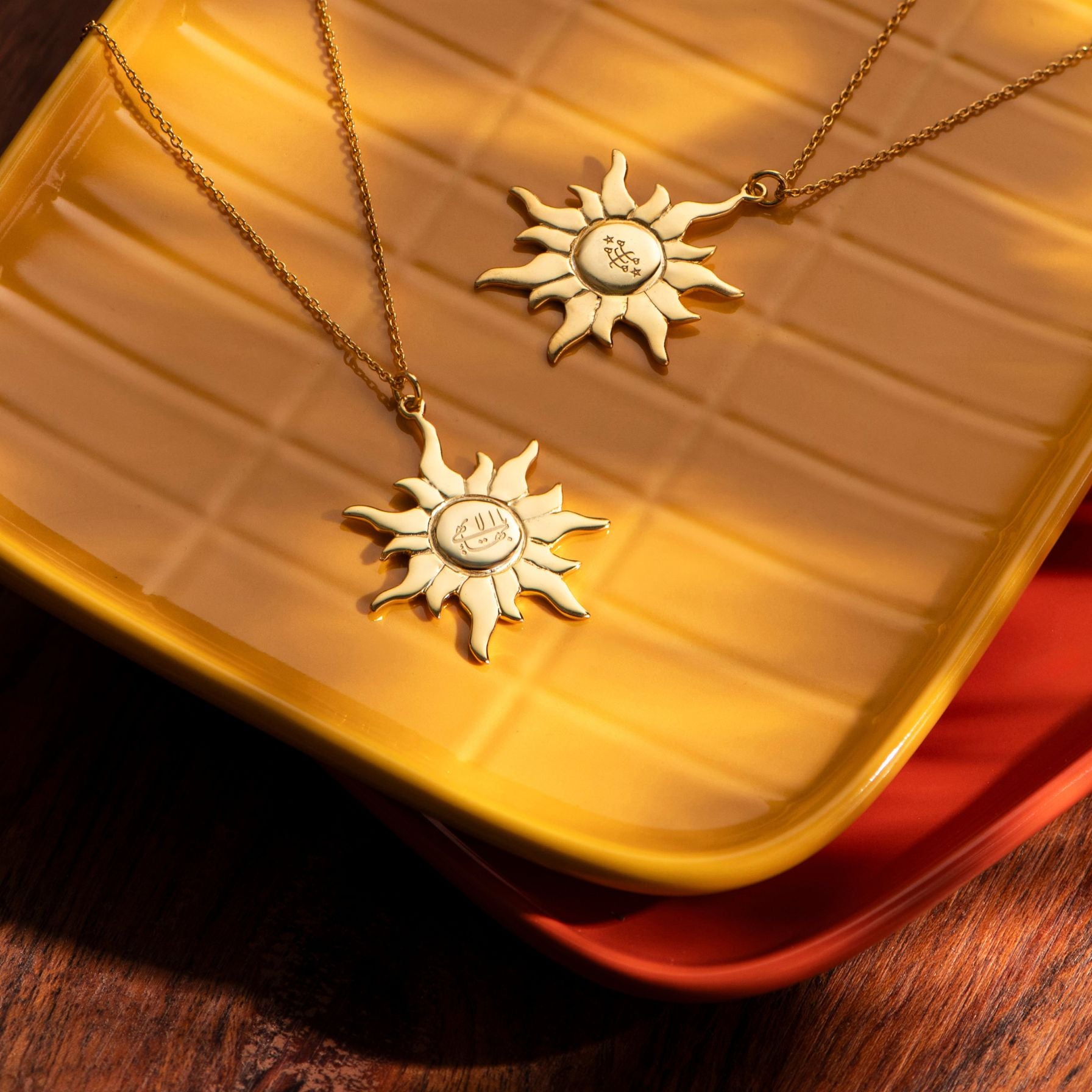 Two gold sun-shaped greatest-name-ringstone-pendants on necklaces against a yellow textured background.