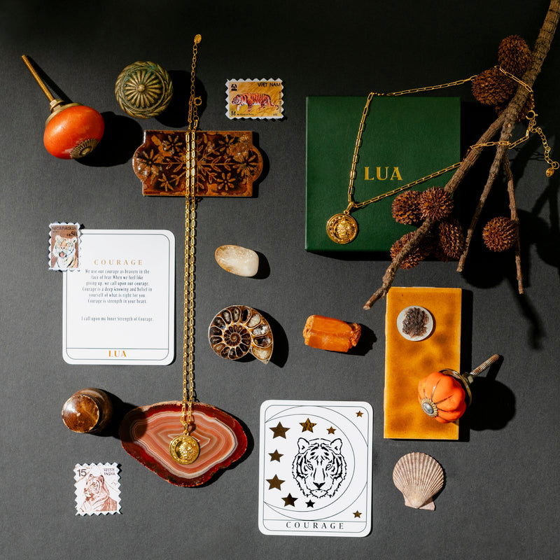 A flat lay image featuring an elegant hue with a gold necklace in 18k Gold Vermeil, a green box, seashells, stamps, dried plants, and cards, including one with a tiger illustration labeled "COURAGE.