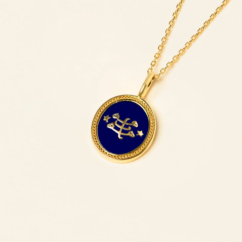 Gold pendant necklace with a navy blue circular enamel charm featuring the ringstone symbol, displayed on a plain beige background.