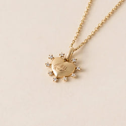 Gold heart-shaped pendant necklace with small cubic zirconia gemstones outlining the heart and featuring a subtle Greatest Name engraving, displayed on a light background.