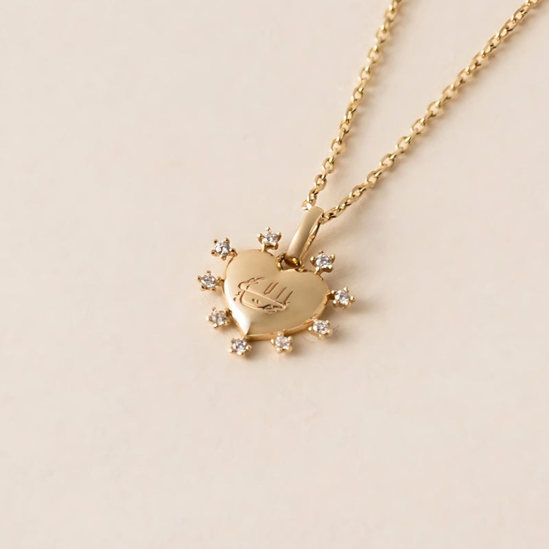 Gold heart-shaped pendant necklace with small cubic zirconia gemstones outlining the heart and featuring a subtle Greatest Name engraving, displayed on a light background.