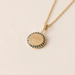 A gold necklace with a round pendant featuring an engraved greatest name symbol, encircled by blue diamonds, displayed against a neutral background.