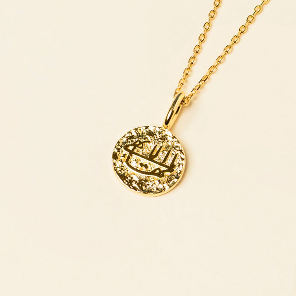 Gold pendant necklace with the Greatest Name in Arabic calligraphy on a pale background.