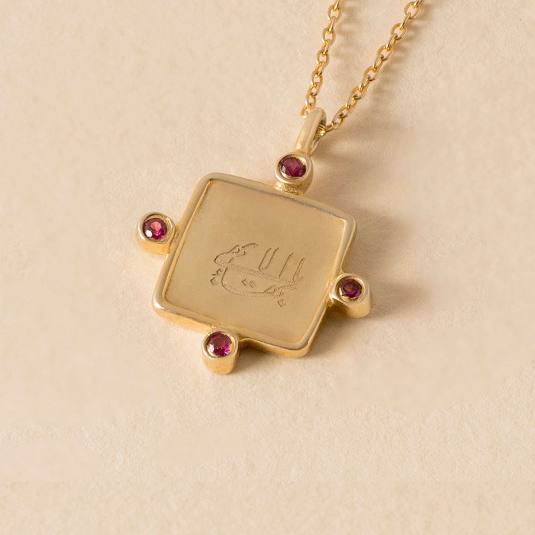 Gold necklace with a square  greatest name pendant featuring engraved text and pink gemstones on a beige background