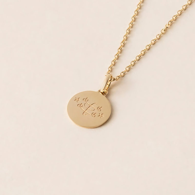 A gold pendant necklace, named the Fortitude Halo Necklace, lies on a light-colored surface. It features a round disc with the Ringstone Symbol engraved in the center.